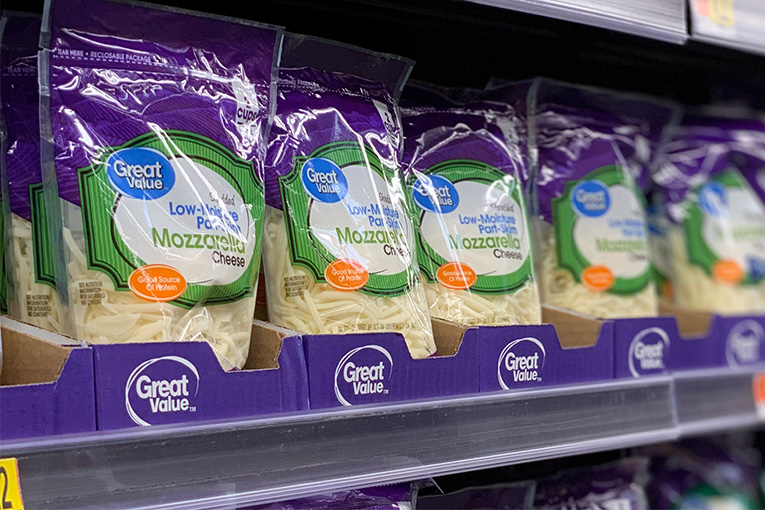 An image of a person holding up a bag of Great Value cheese displayed in a Delkor Cabrio Case with a wall of different cheese packages behind it